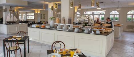A breakfast buffet area with a central island, dining tables, chairs, and a view of the outdoors through large windows.