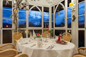 A dining area with a round table set for a meal, surrounded by wicker chairs, and large windows with decorative elements.