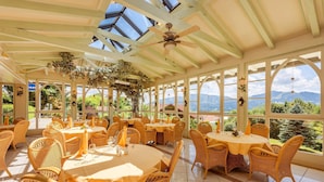 A sunlit dining area with wicker furniture, large windows, and a view of mountains.