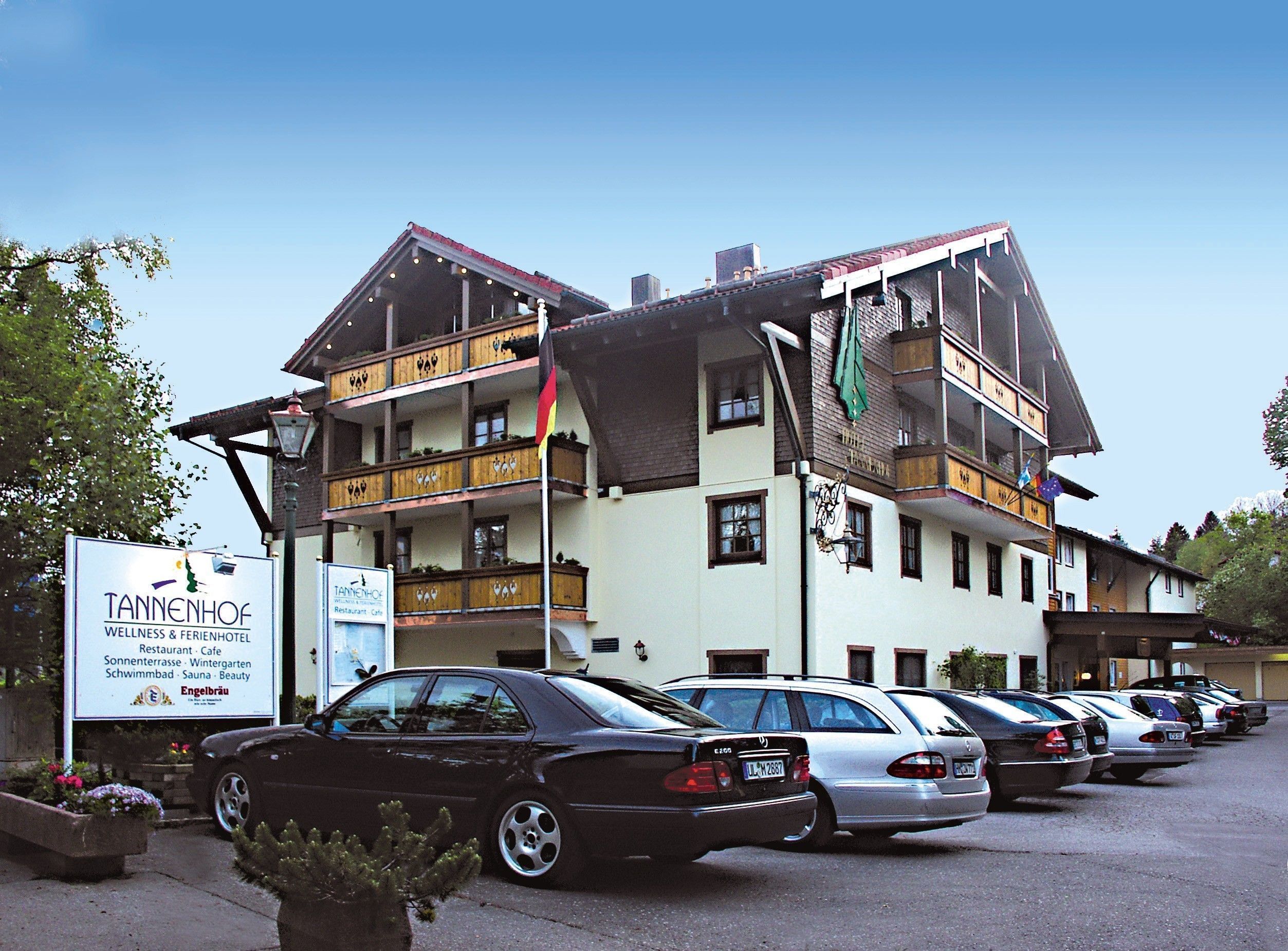 Ein Hotel mit mehreren Balkonen, ein Parkplatz mit einigen Autos und ein Schild, das Wellness- und Restaurantservice bewirbt.