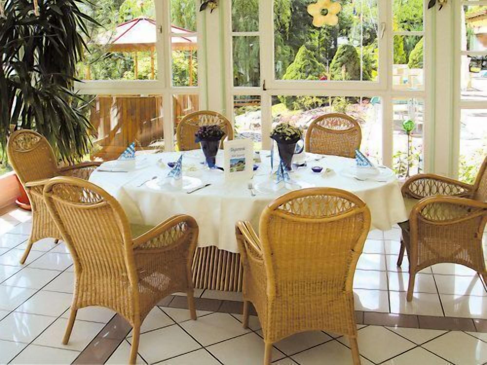 A dining area with wicker chairs and a round table set for six, surrounded by large windows.