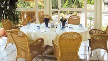 A dining area with wicker chairs and a round table set for six, surrounded by large windows.