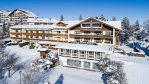 A large, multi-story hotel with a ski lift visible in the background, surrounded by snow-covered trees and a clear blue sky.