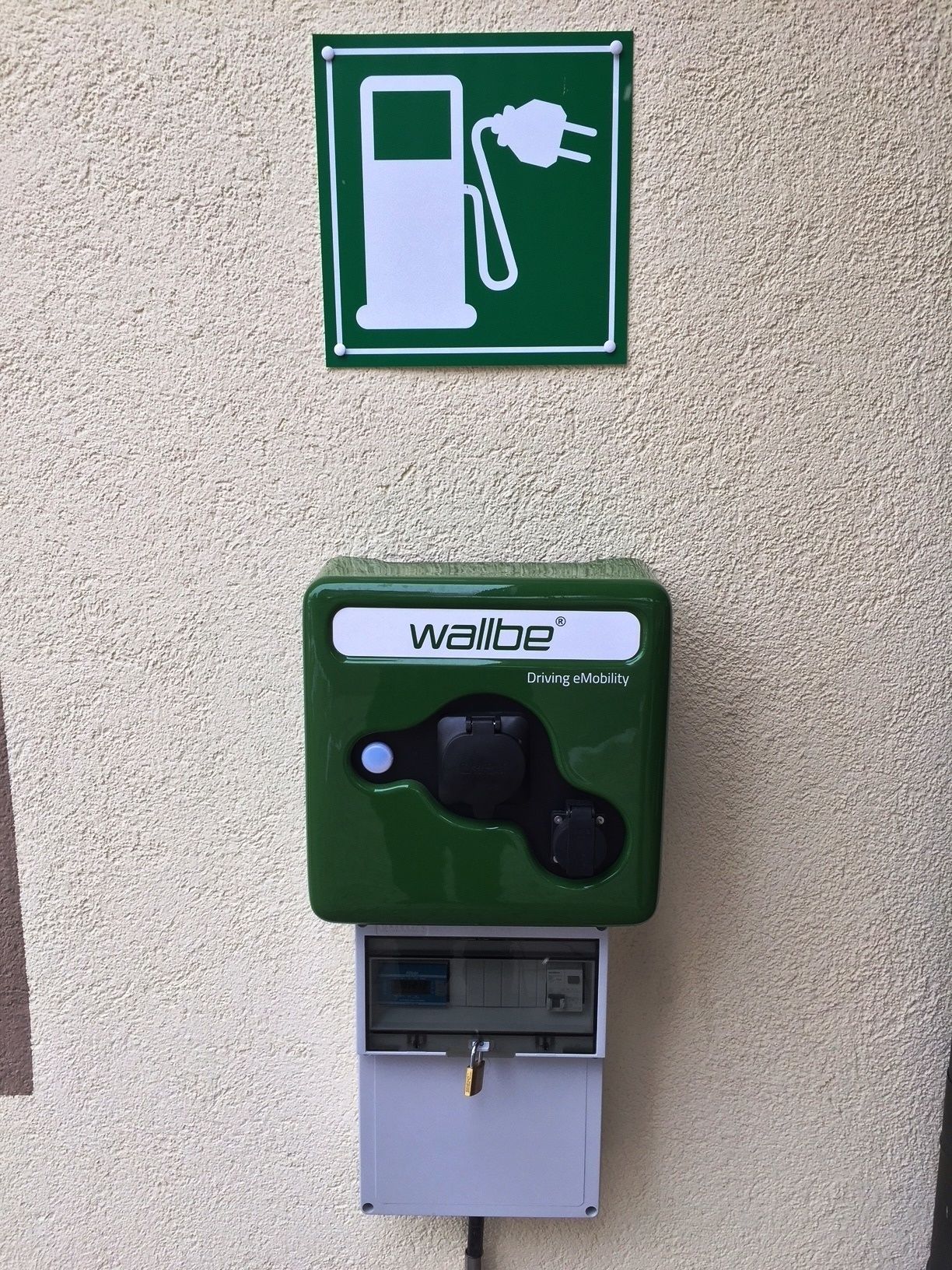 A green electric vehicle charging station with a plug and a charging cable, mounted on a textured wall.