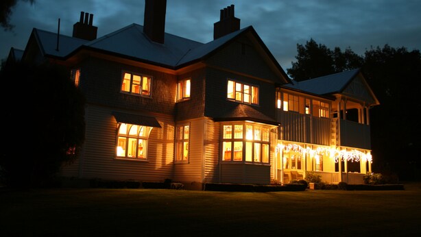 A large, two-story house with multiple windows and a porch, illuminated by warm interior lighting.