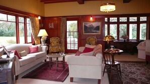 A cozy living room with a sofa, armchairs, a coffee table, and a chandelier. There are large windows with curtains, a painting on the wall, and a view of greenery outside.