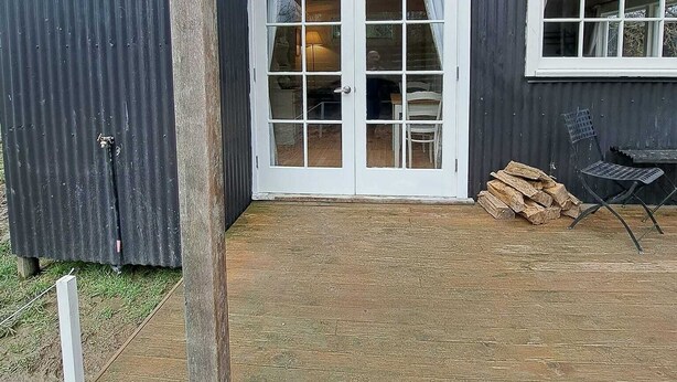 A covered patio area with a wooden deck, a white door with glass panels, and a metal chair. - Gunyah Country Estate (Windwhistle)