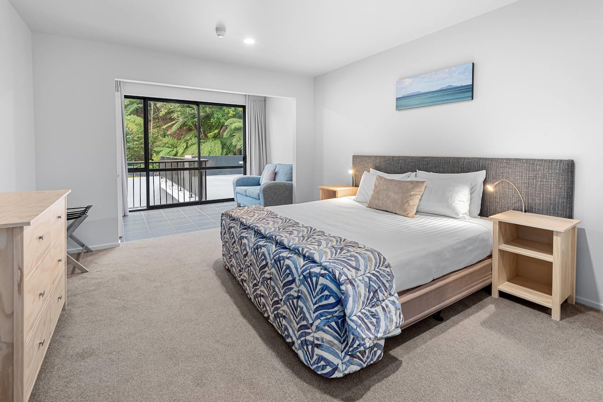 A bedroom with a large bed, a nightstand, a dresser, and a balcony with a view of greenery.