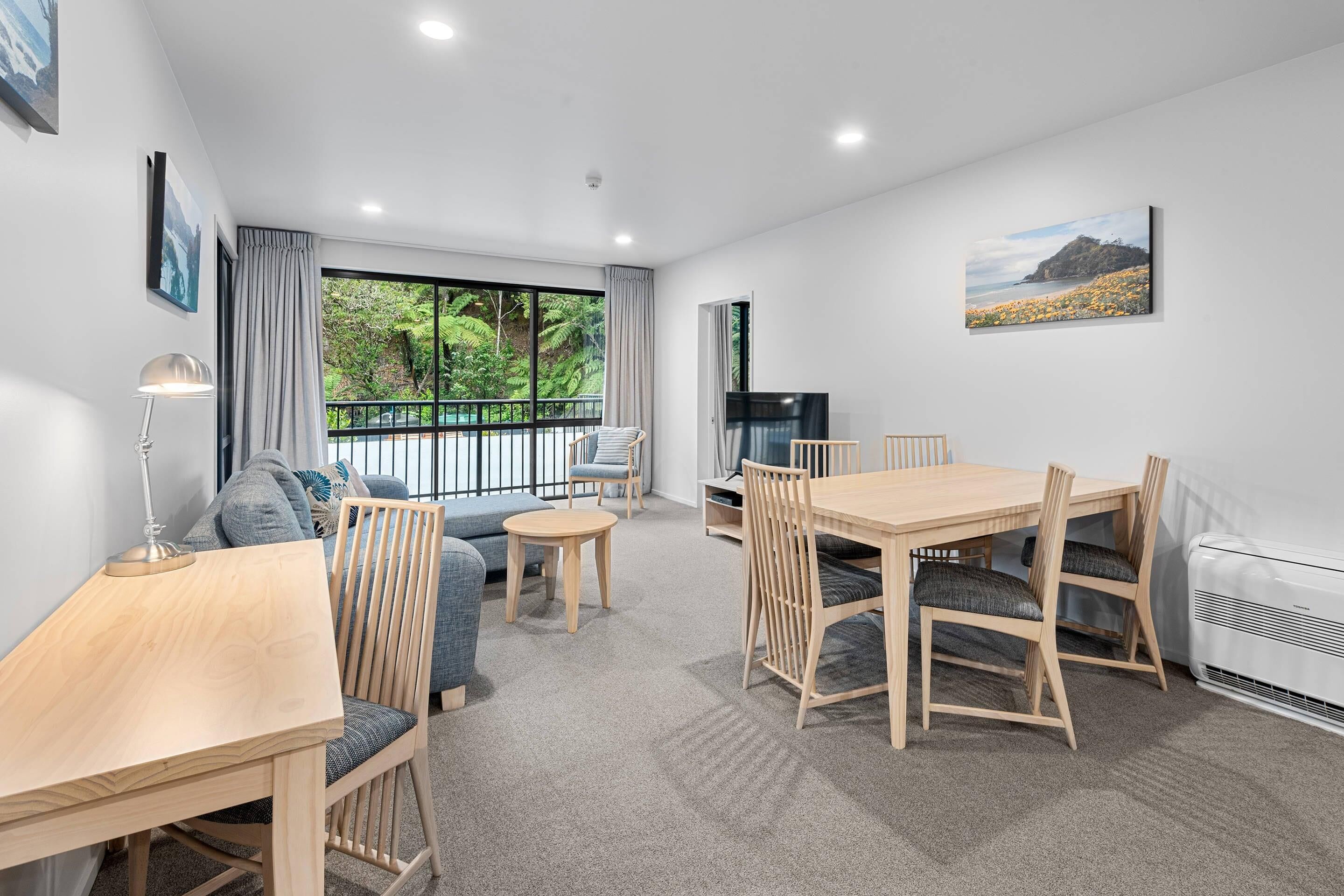 A modern living room with a dining area, large windows, and a view of greenery.