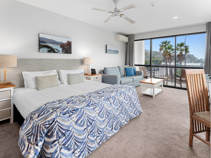 A hotel room with a bed, a sofa, a desk, a chair, a balcony with a view, and a ceiling fan.