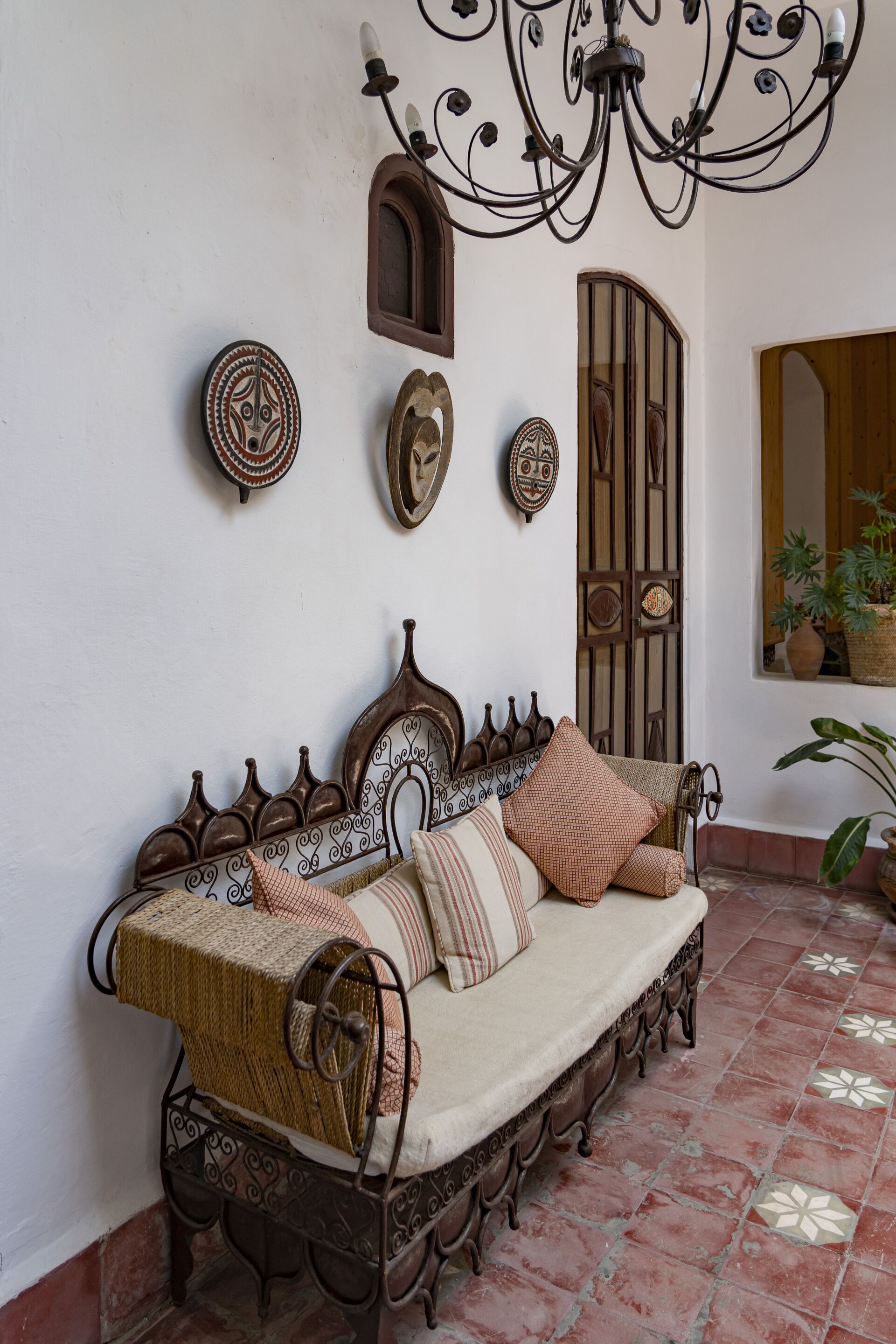 A traditional wooden bench with a woven seat and cushions, a decorative wall with a chandelier, and a tiled floor.