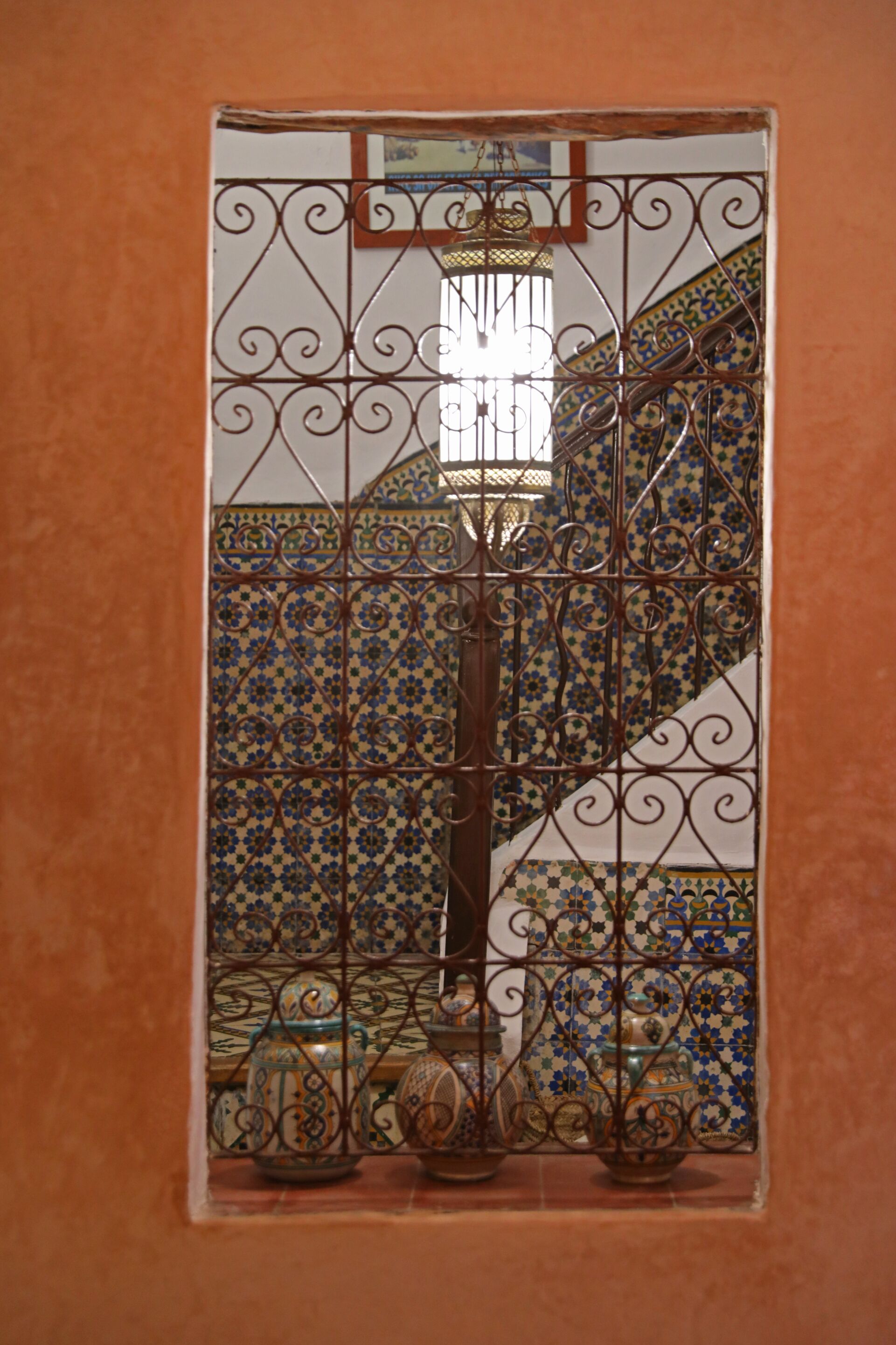 A view through a window with ornate metal bars, a tiled wall with geometric patterns, and a staircase with a decorative railing.