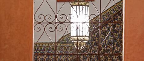 A view through a window with ornate metal bars, a tiled wall with geometric patterns, and a staircase with a decorative railing.