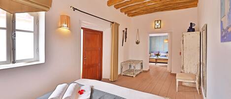 A bedroom with a bed, a wooden ceiling, a window, a door, and a view of another room.