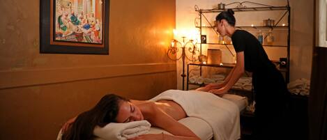 A massage therapy room with a client lying on a massage table, a therapist attending to the client, and a wall-mounted shelf with towels and bottles.