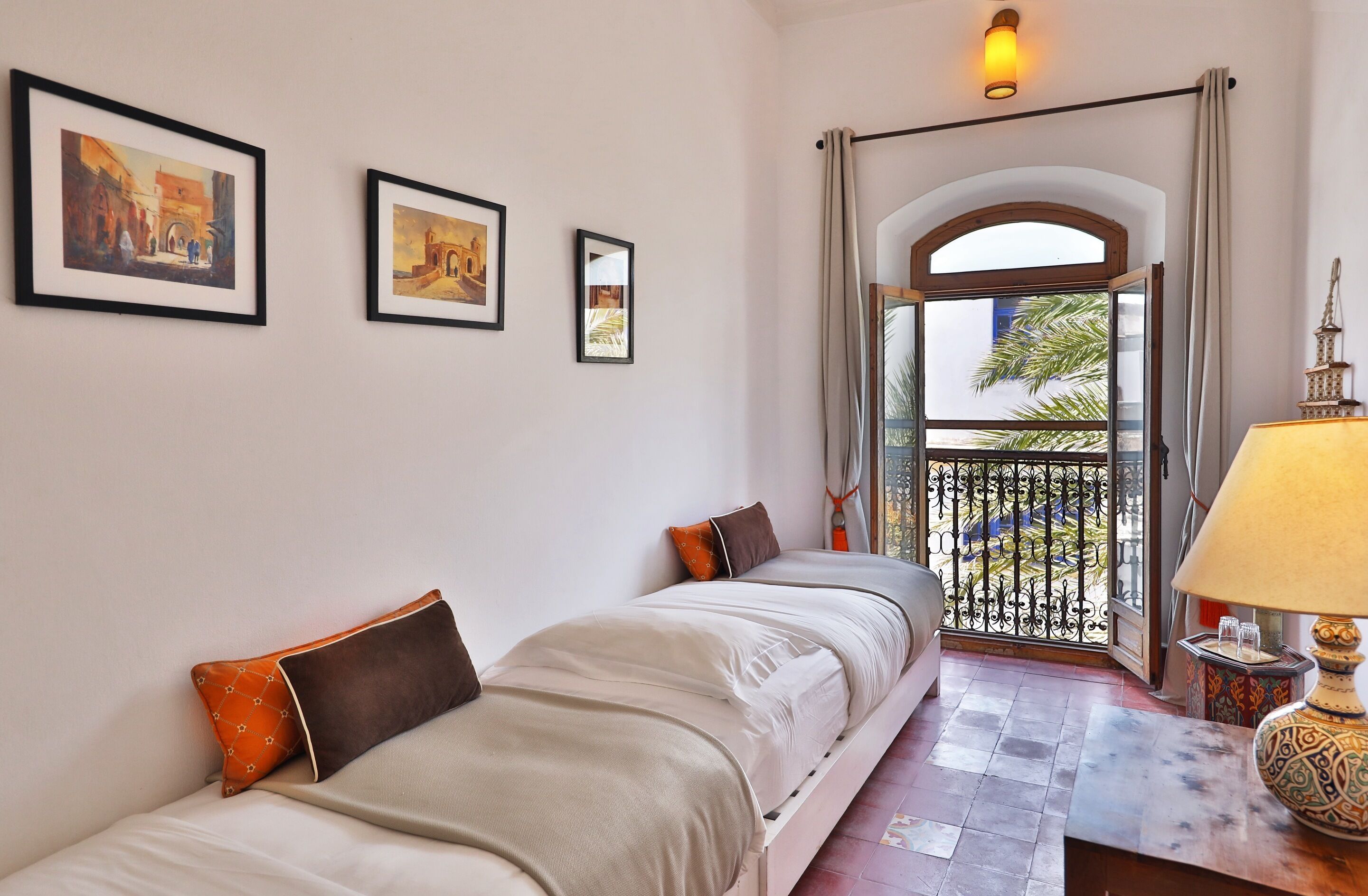 Habitación de hotel con dos camas, un balcón con barandilla de hierro forjado y cuadros en la pared.