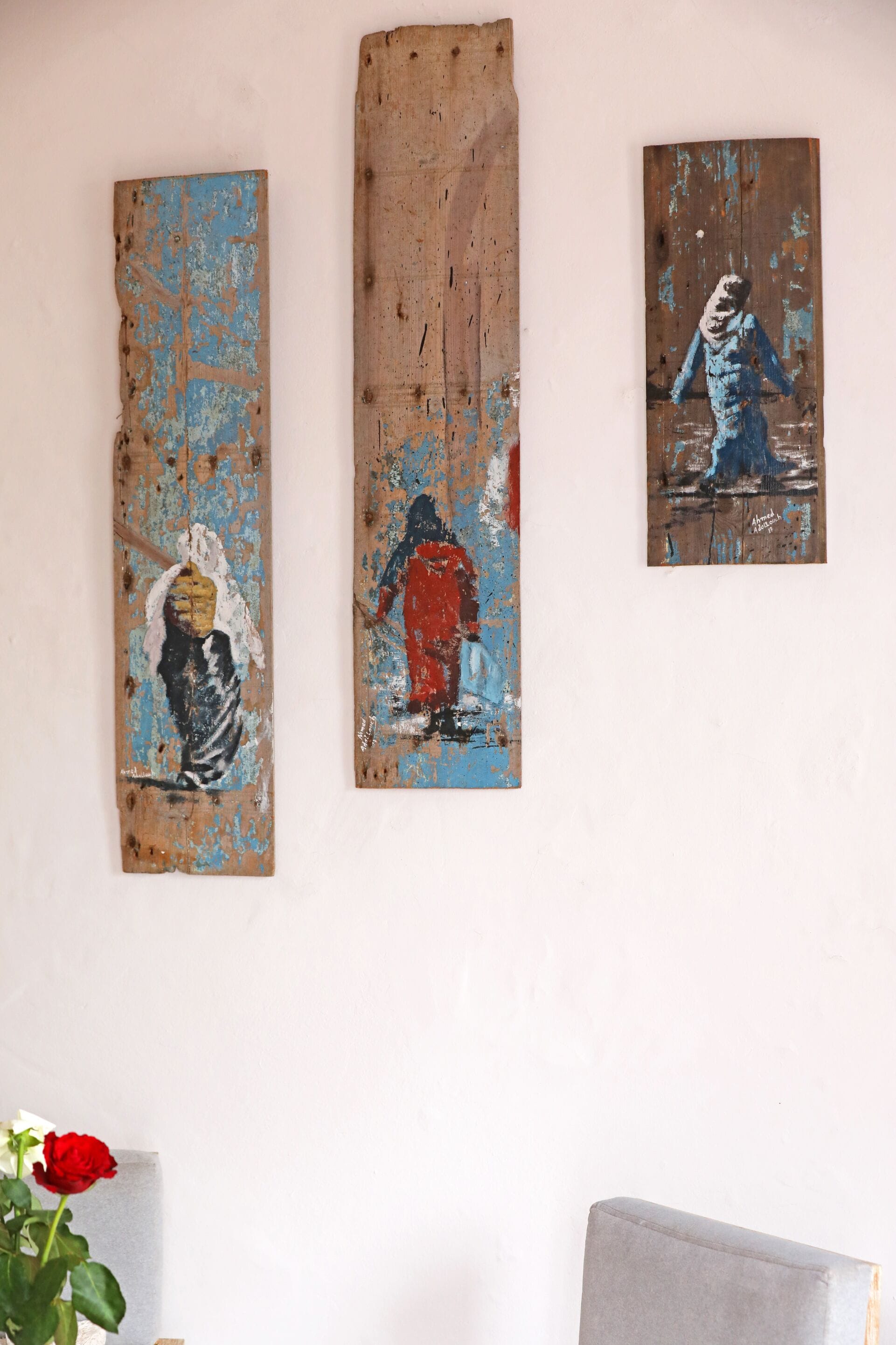 Three artworks on a wall, a vase with a red rose, and a grey chair.