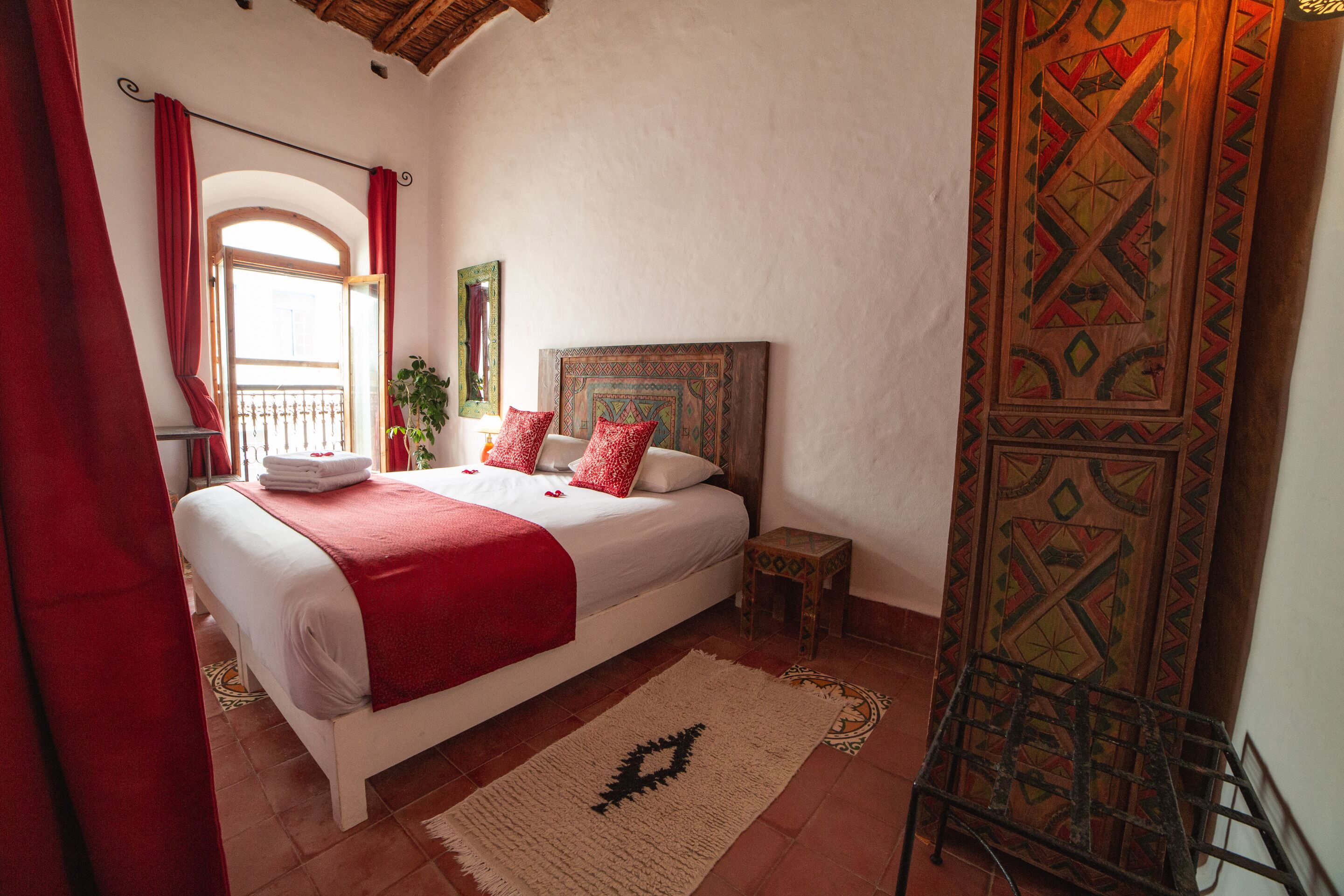 A bedroom with a bed, a nightstand, a red curtain, a window, and a patterned wall.