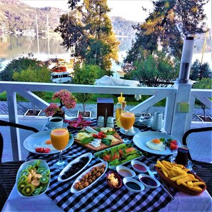 A table set with breakfast items, including fruits, eggs, and beverages, overlooking a lake with boats and a forested hill in the background. - Levent Hotel (Fethiye)