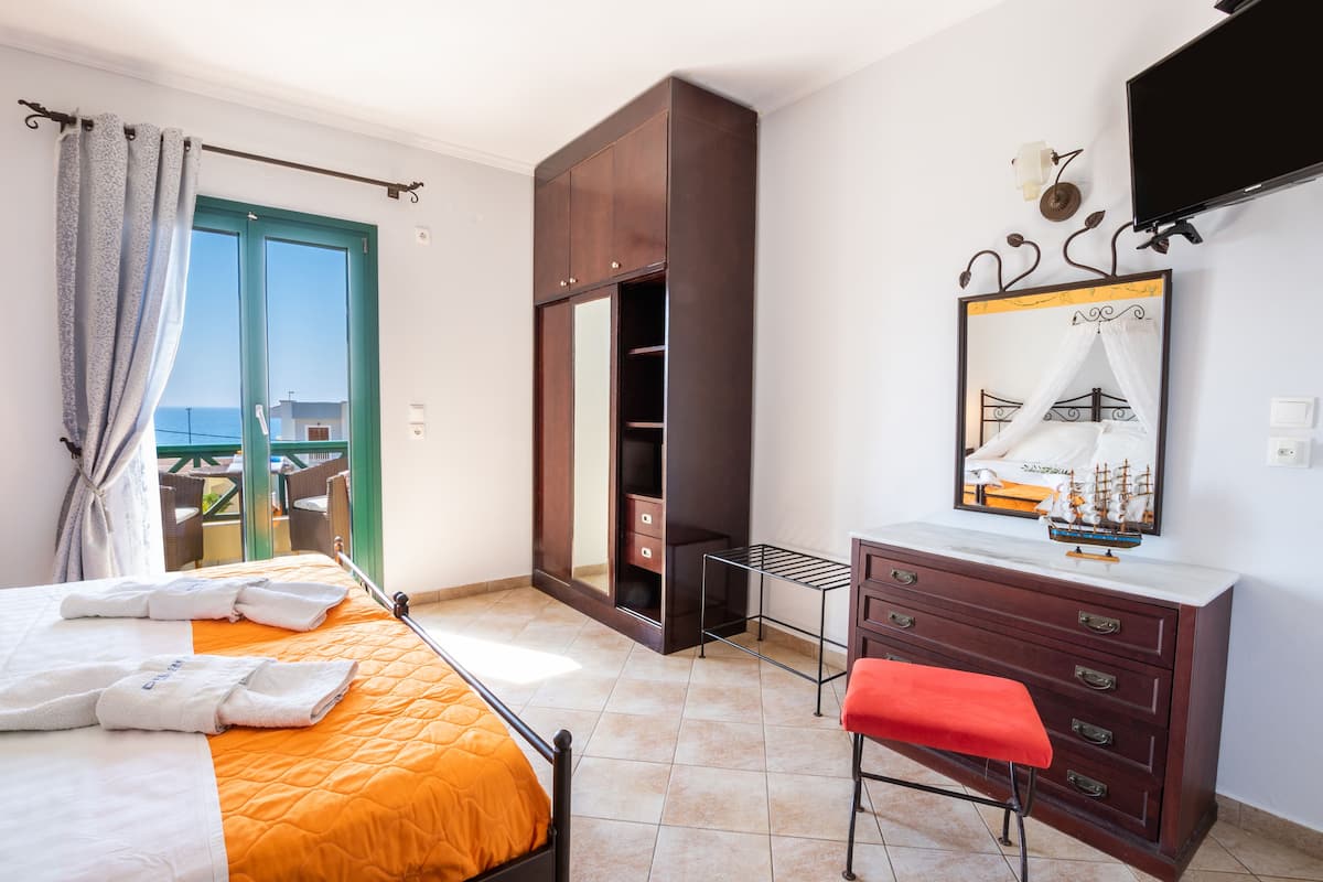 A hotel room with a bed, a wardrobe, a desk, a chair, a bench, a mirror, and a television.
