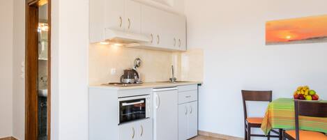 Una cocina compacta con armarios blancos, horno integrado y cafetera. Se ve una mesa de comedor con un frutero.