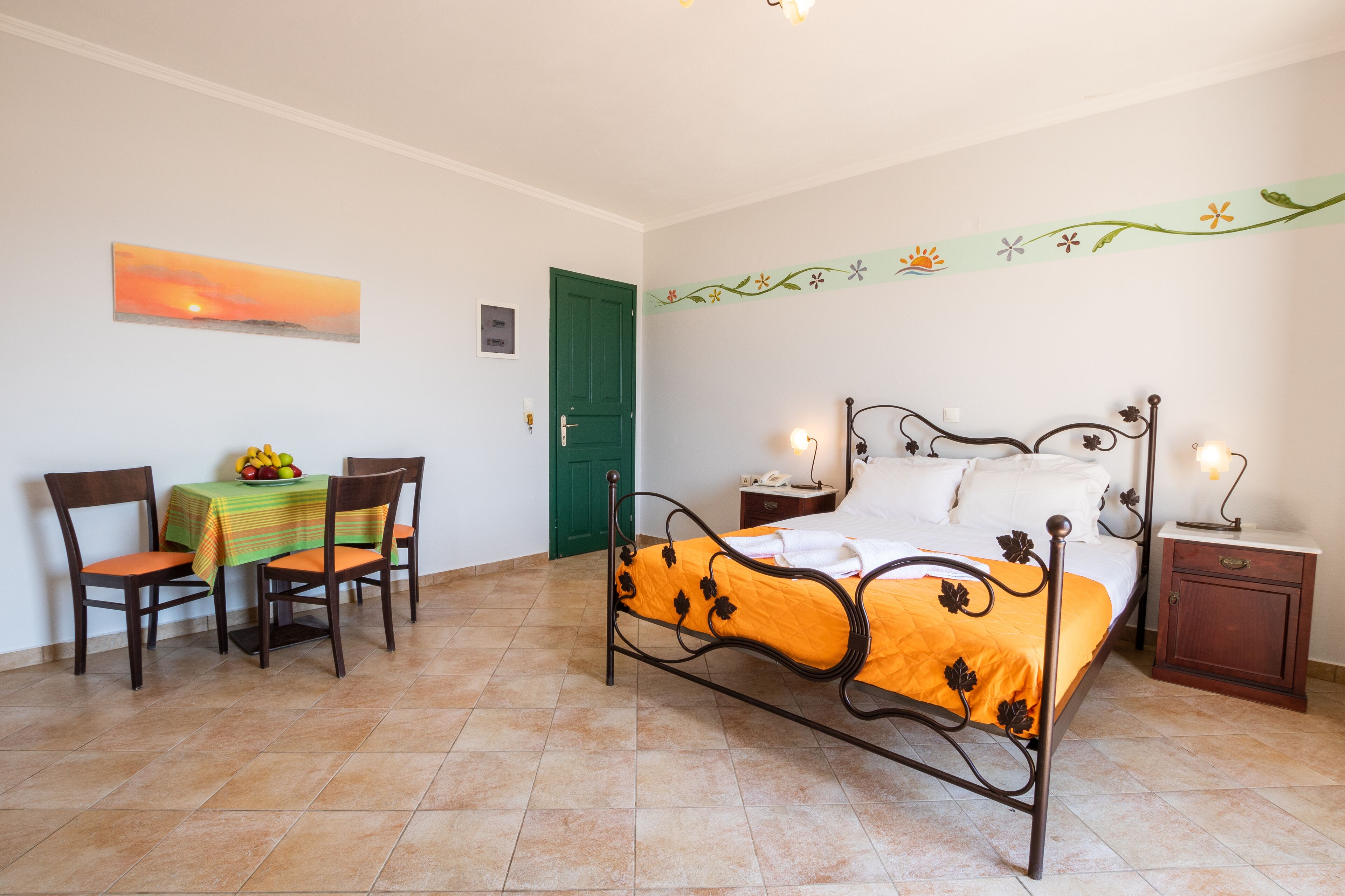 Ein Schlafzimmer mit einem Bett, einem Nachttisch, einem Beistelltisch, einem Stuhl, einem Tisch mit Obstschale und einer grünen Tür.