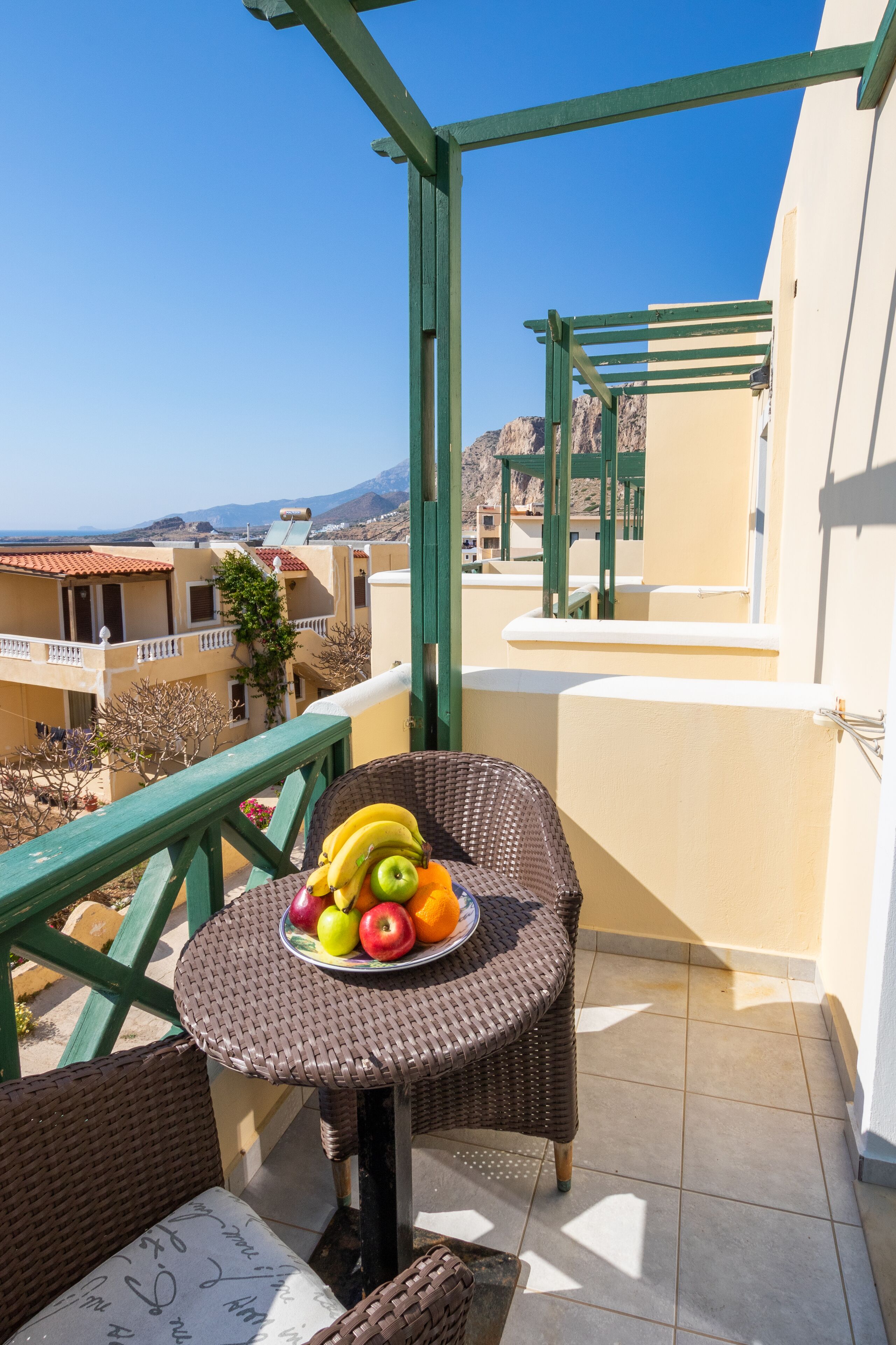 Un balcón con mobiliario de mimbre, un frutero y vistas a edificios y montañas.