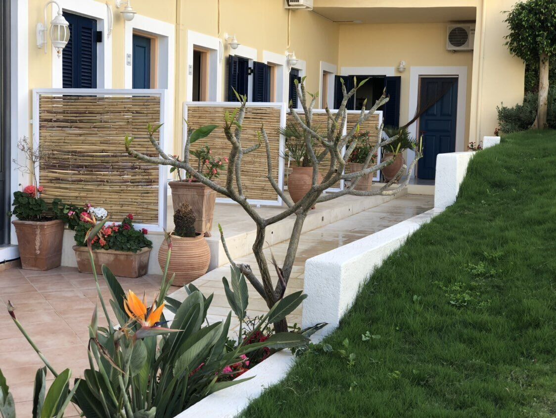 Fila de habitaciones de hotel con paredes blancas y contraventanas oscuras, cada una con un pequeño balcón y plantas en macetas.