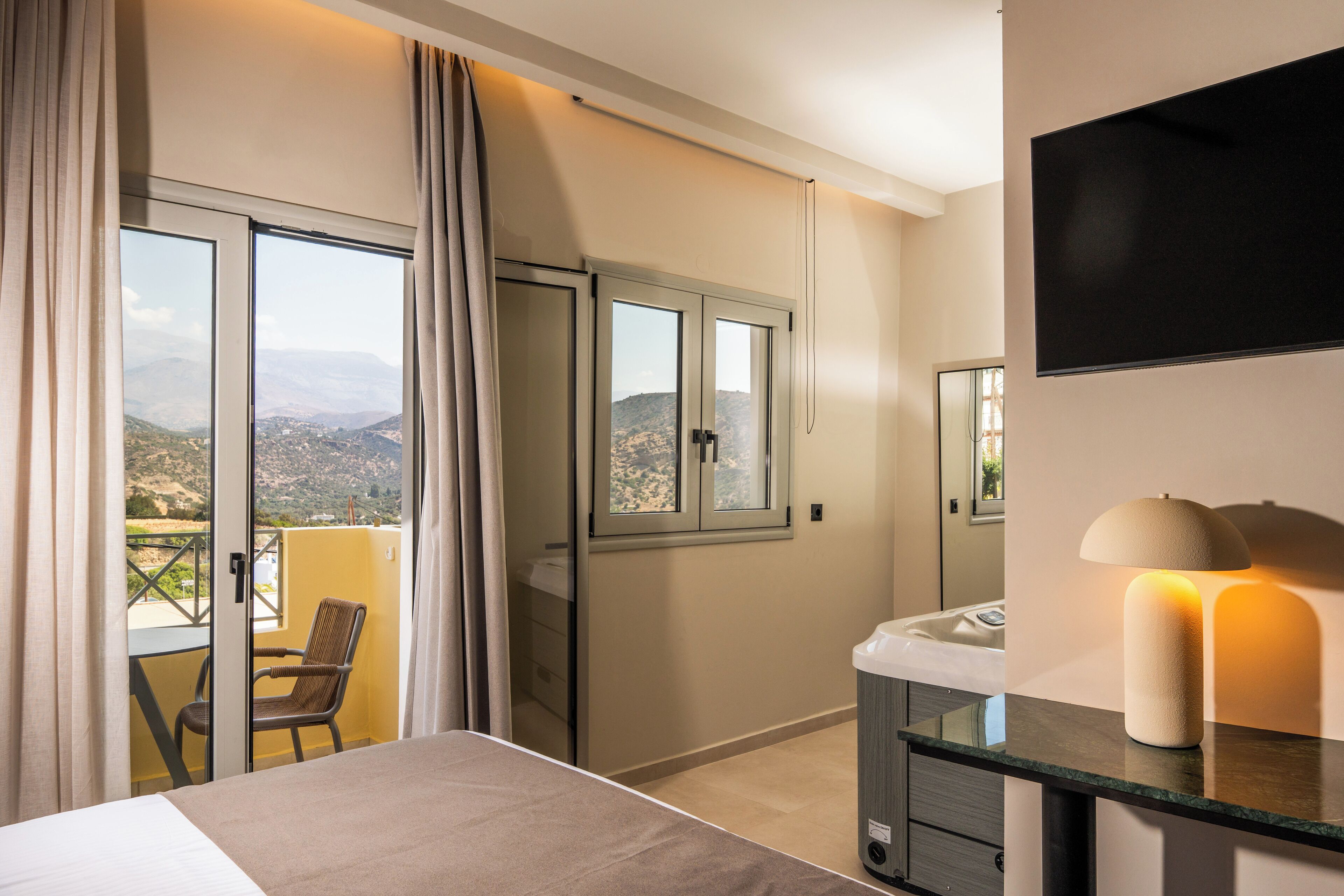 A hotel room with a bed, a chair, a balcony with curtains, a TV, and a lamp.