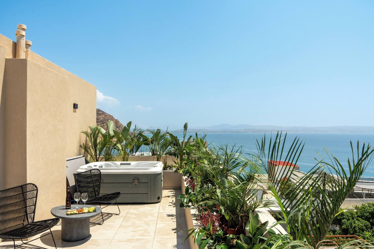 A terrace with a hot tub, seating, and plants, overlooking a coastal view.