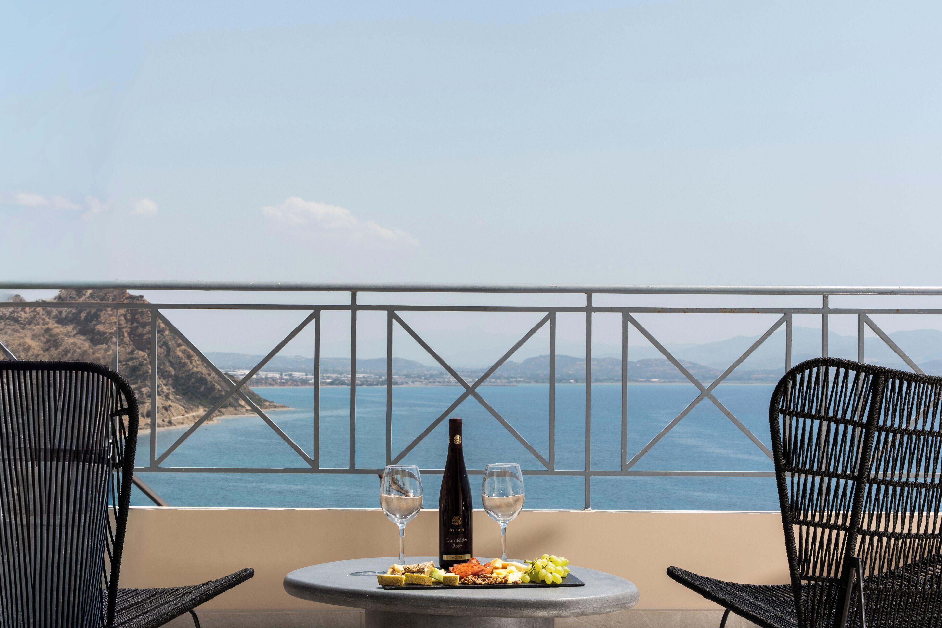 A table with a bottle of wine, two glasses, and a plate of food on a balcony overlooking a coastal view.