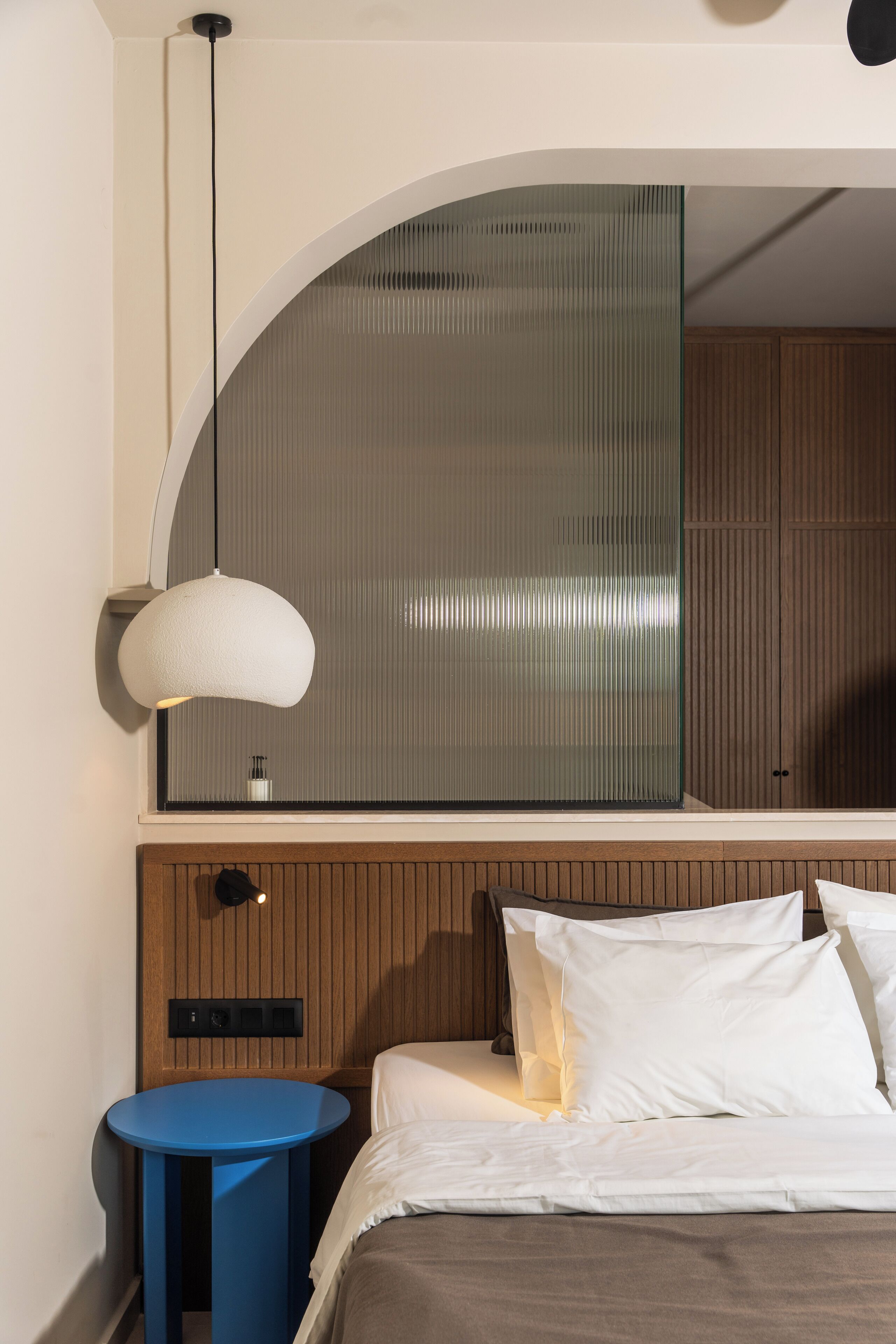 Un dormitorio con cama, una mesita de noche azul, una lámpara colgante blanca y un cabecero de madera.