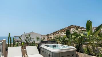 A rooftop terrace with a hot tub, lounge chairs, and a view of the sea and hills.