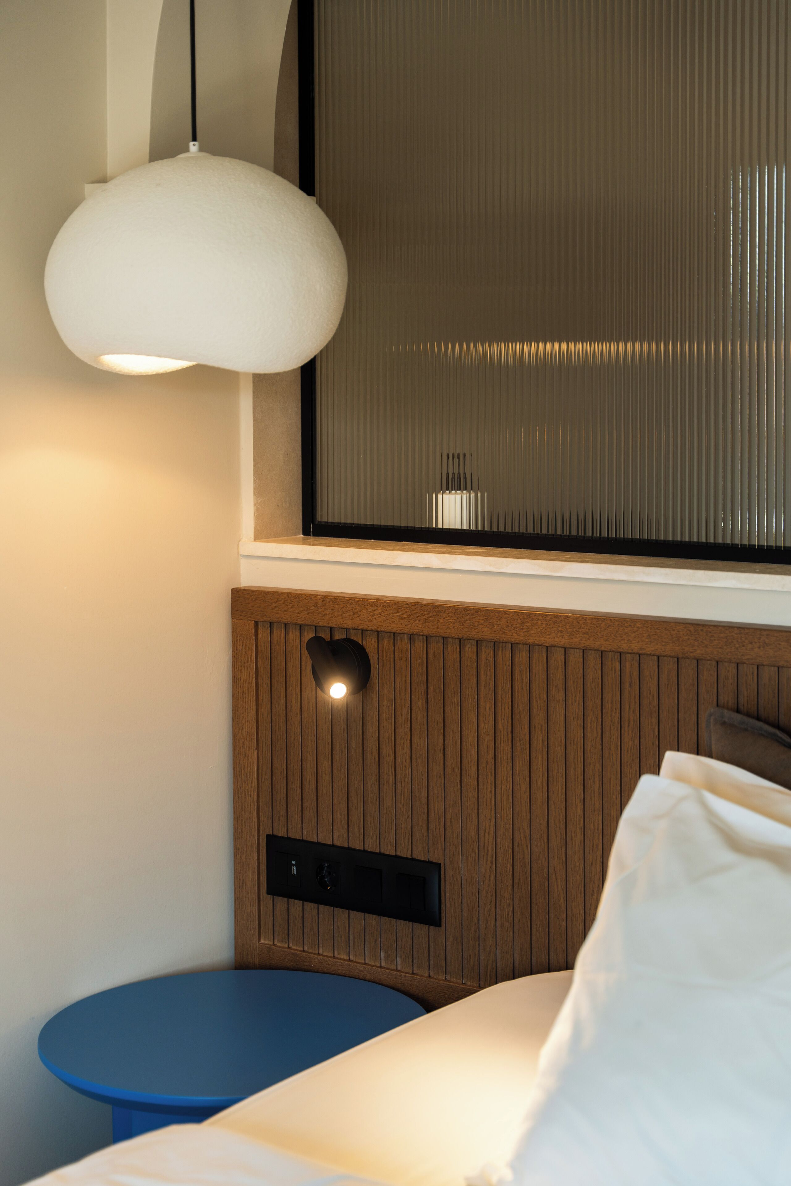 A hotel room with a bed, a bedside table, a wall-mounted light, and a window with blinds.