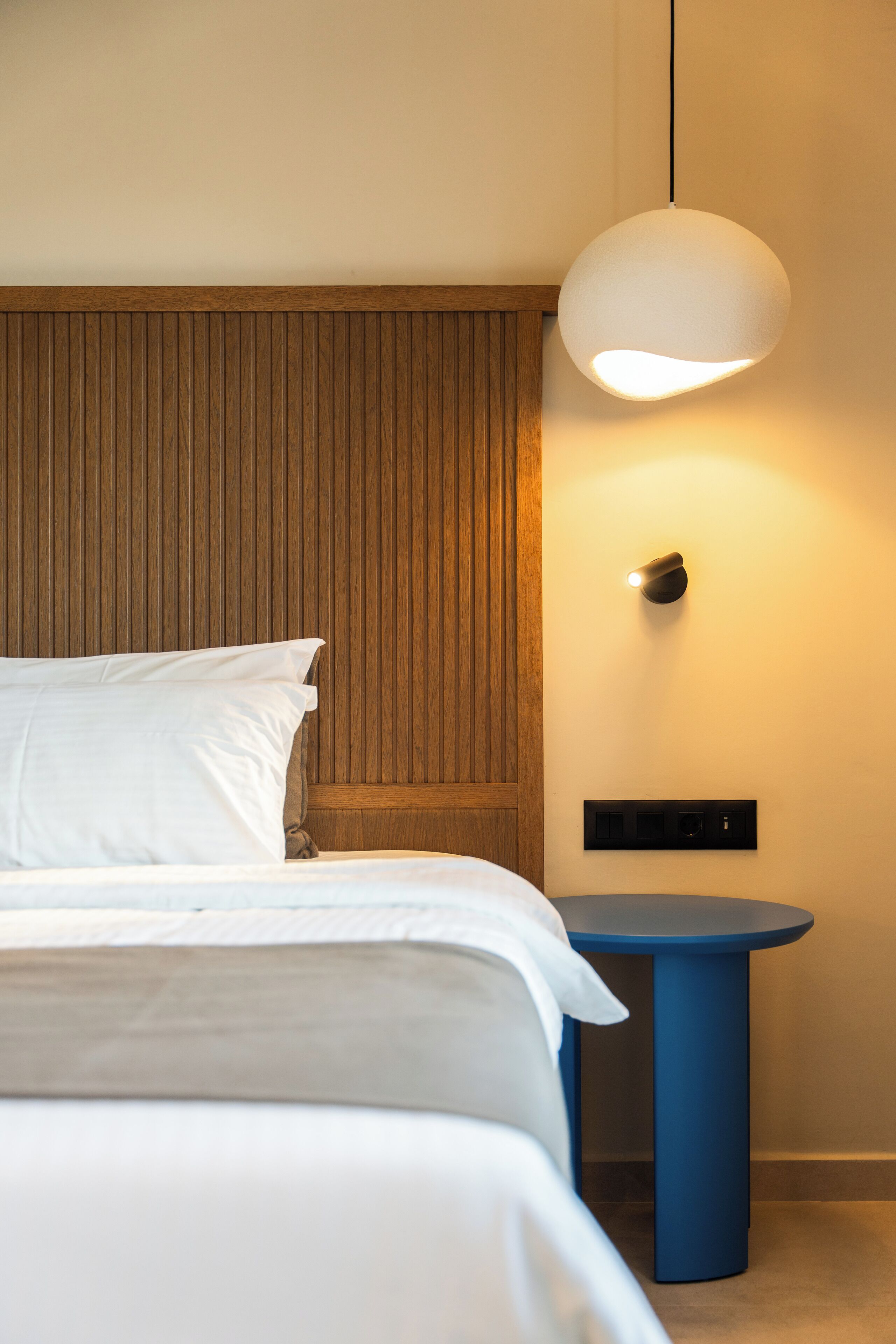 A neatly made bed with white linens, a wooden headboard, a round blue side table, and a hanging light fixture.