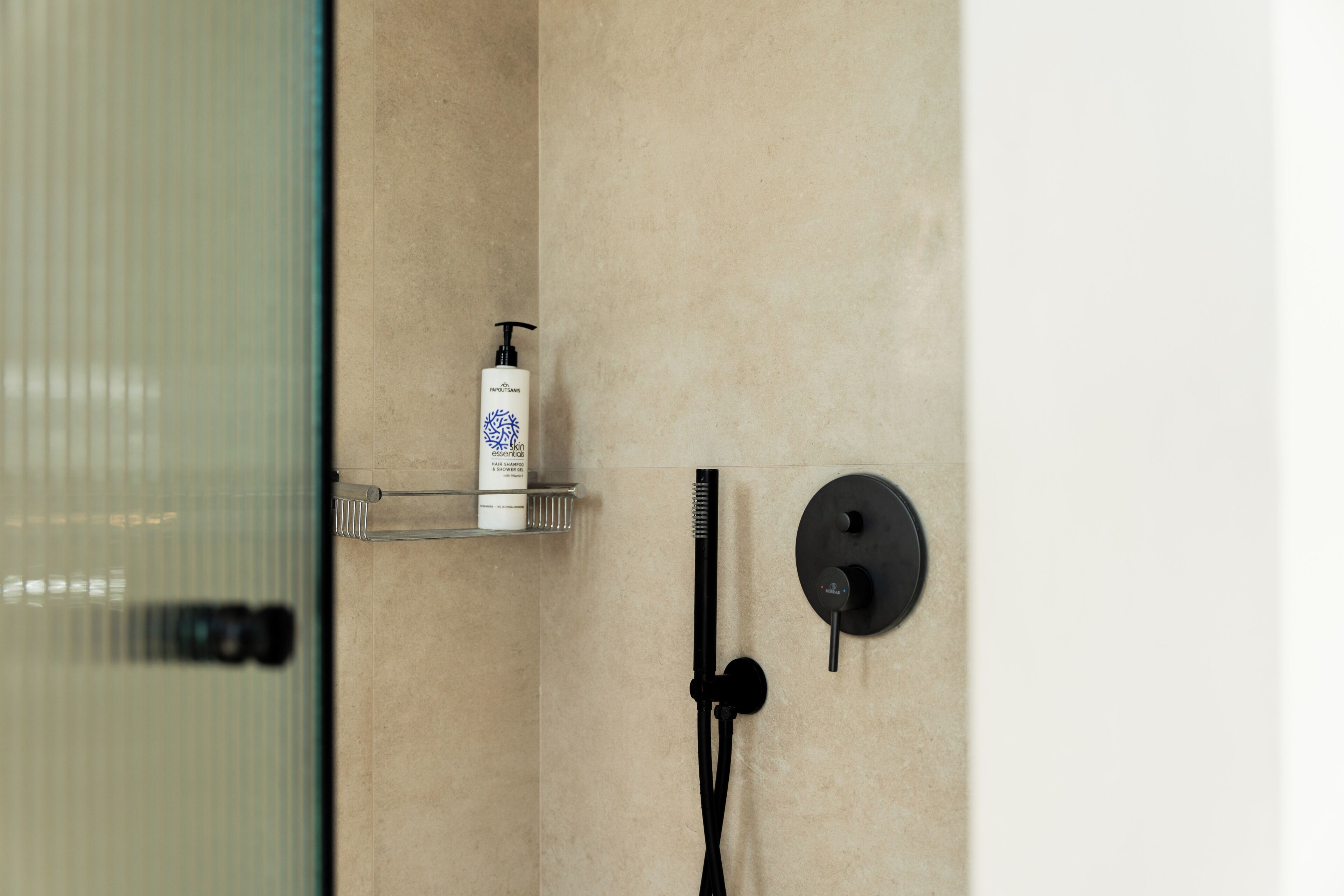A bathroom with a glass shower enclosure, a black handheld showerhead, and a white bottle of shampoo on a shelf.