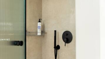 Un baño con cabina de ducha de vidrio, un cabezal de ducha manual negro y una botella blanca de champú en un estante.