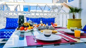 A breakfast table with a variety of fruits, coffee, and orange juice, set on a balcony with blue cushions and a yellow pot. - Dar Nafoura Mogador (Essaouira)