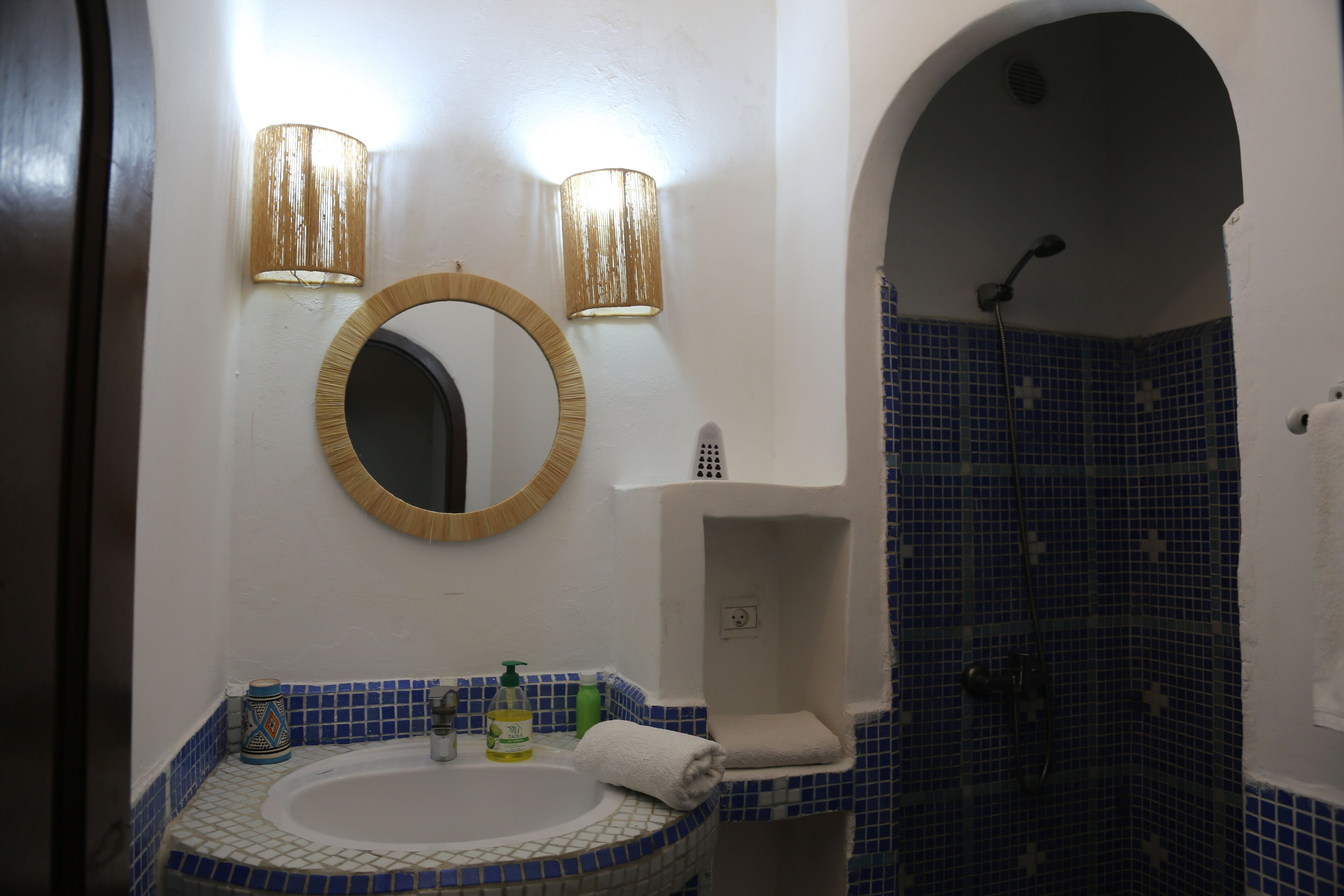 Une salle de bain avec un miroir rond, deux appliques murales, un lavabo et un espace douche agrémenté de carreaux de mosaïque bleus.