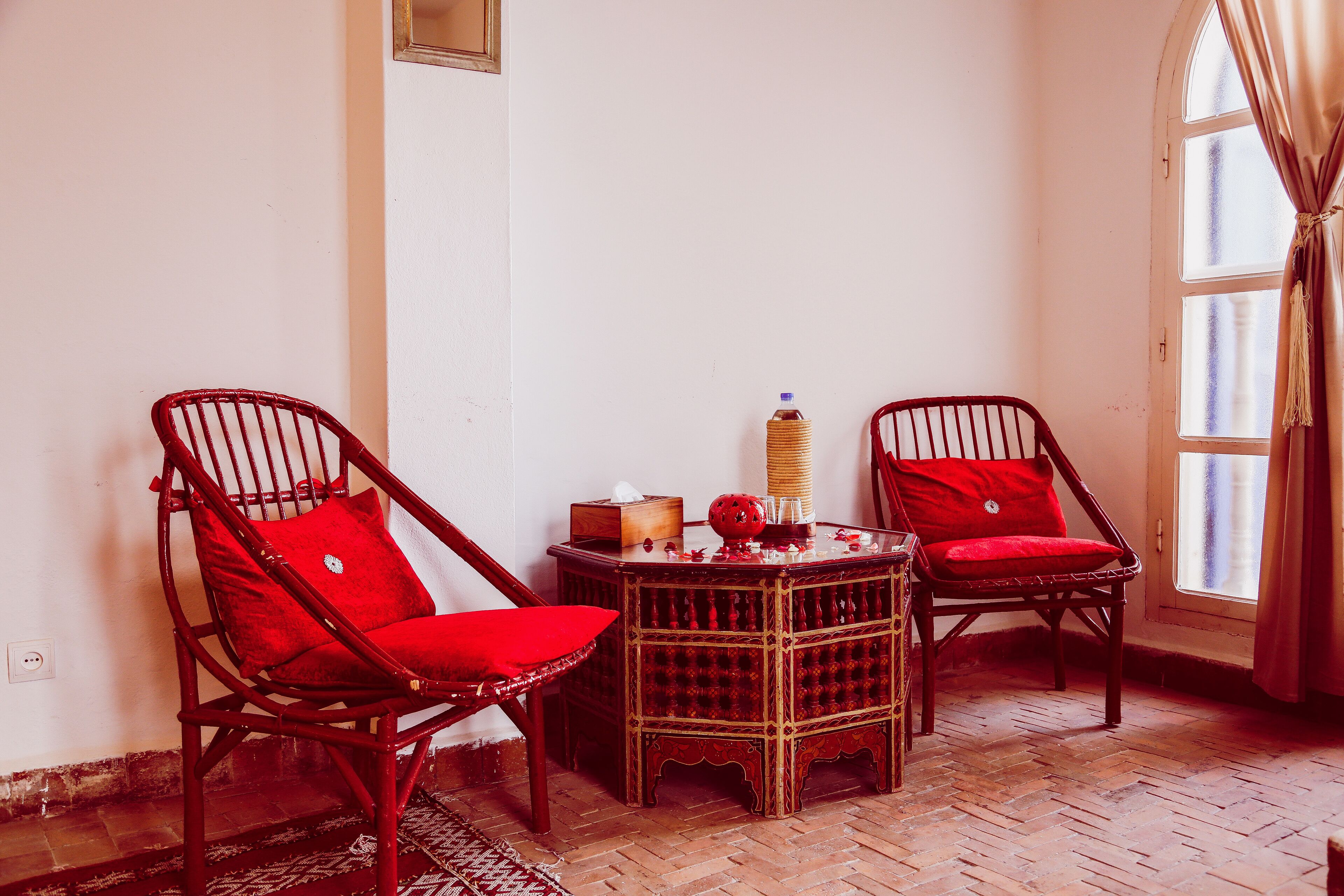 Una habitación con dos sillas de mimbre y cojines rojos, una mesita de madera con una lámpara y una ventana con cortinas.