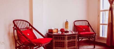 A room with two wicker chairs with red cushions, a small wooden table with a lamp, and a window with curtains.
