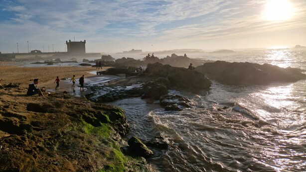 A coastal scene with rocky terrain, a castle-like structure, and the ocean. - Dar Nafoura Mogador (Essaouira)