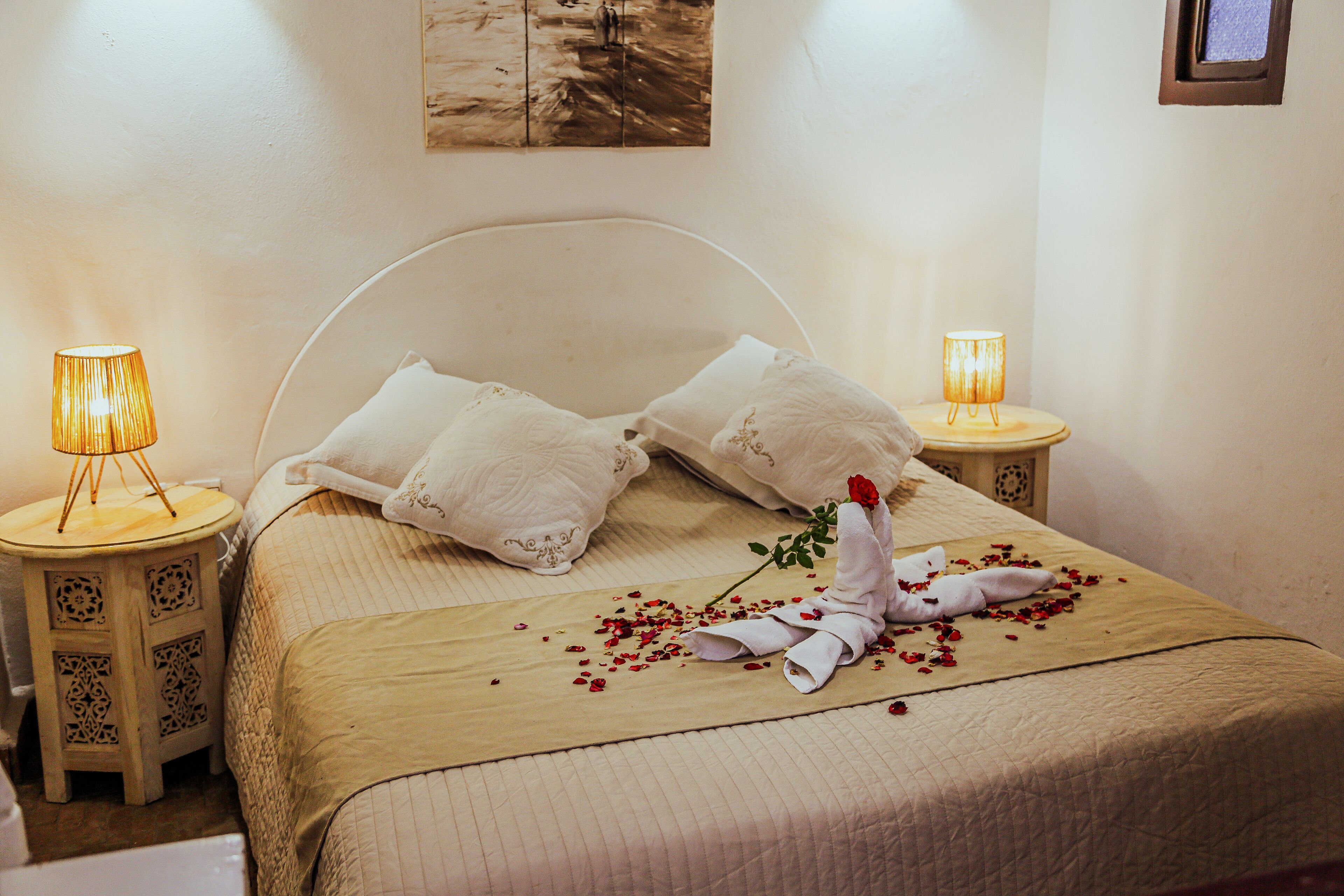 A neatly made bed with pillows, a rose, and scattered petals. There are two bedside lamps on wooden nightstands.