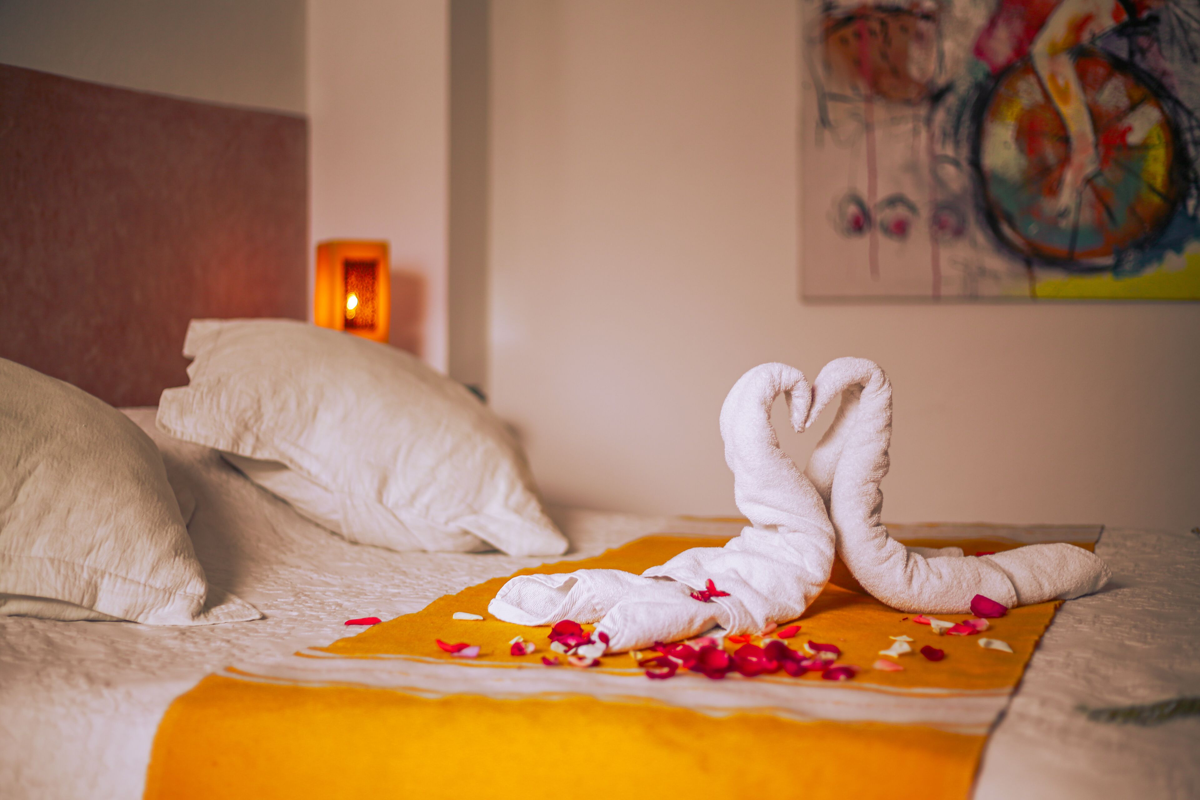 A bed with white pillows and a yellow blanket, a towel arranged in a heart shape, and rose petals scattered on the blanket.