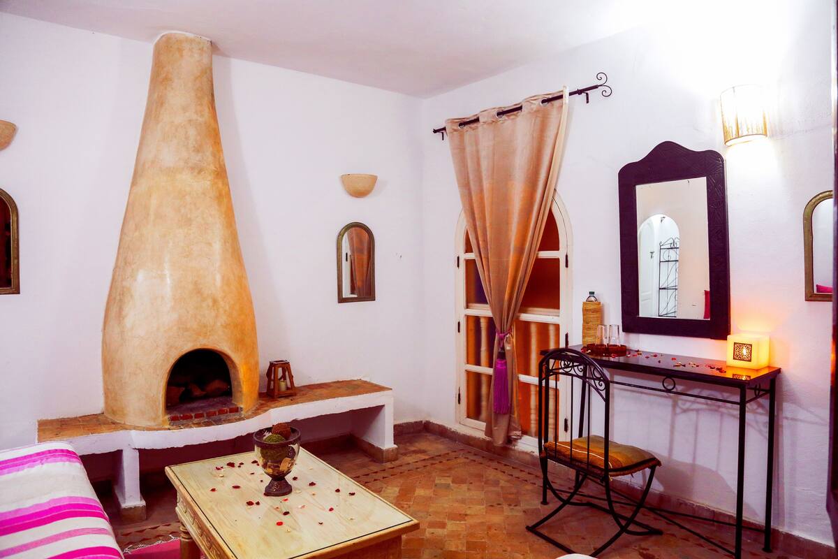 A cozy room with a clay oven, a wooden table with rose petals, a chair, a mirror, and a lamp.