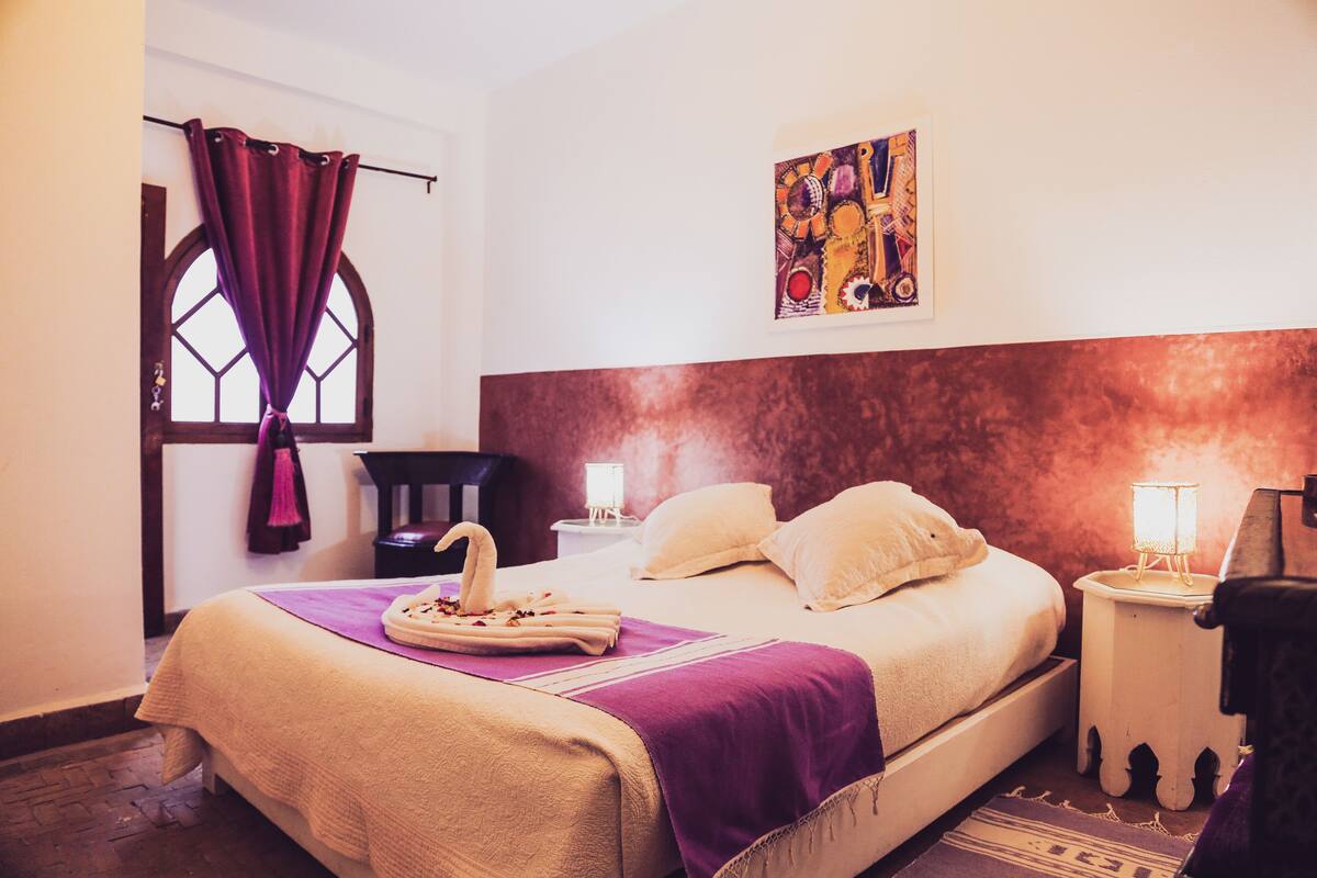 A hotel room with a large bed, a window with curtains, a nightstand with a lamp, and a painting on the wall.