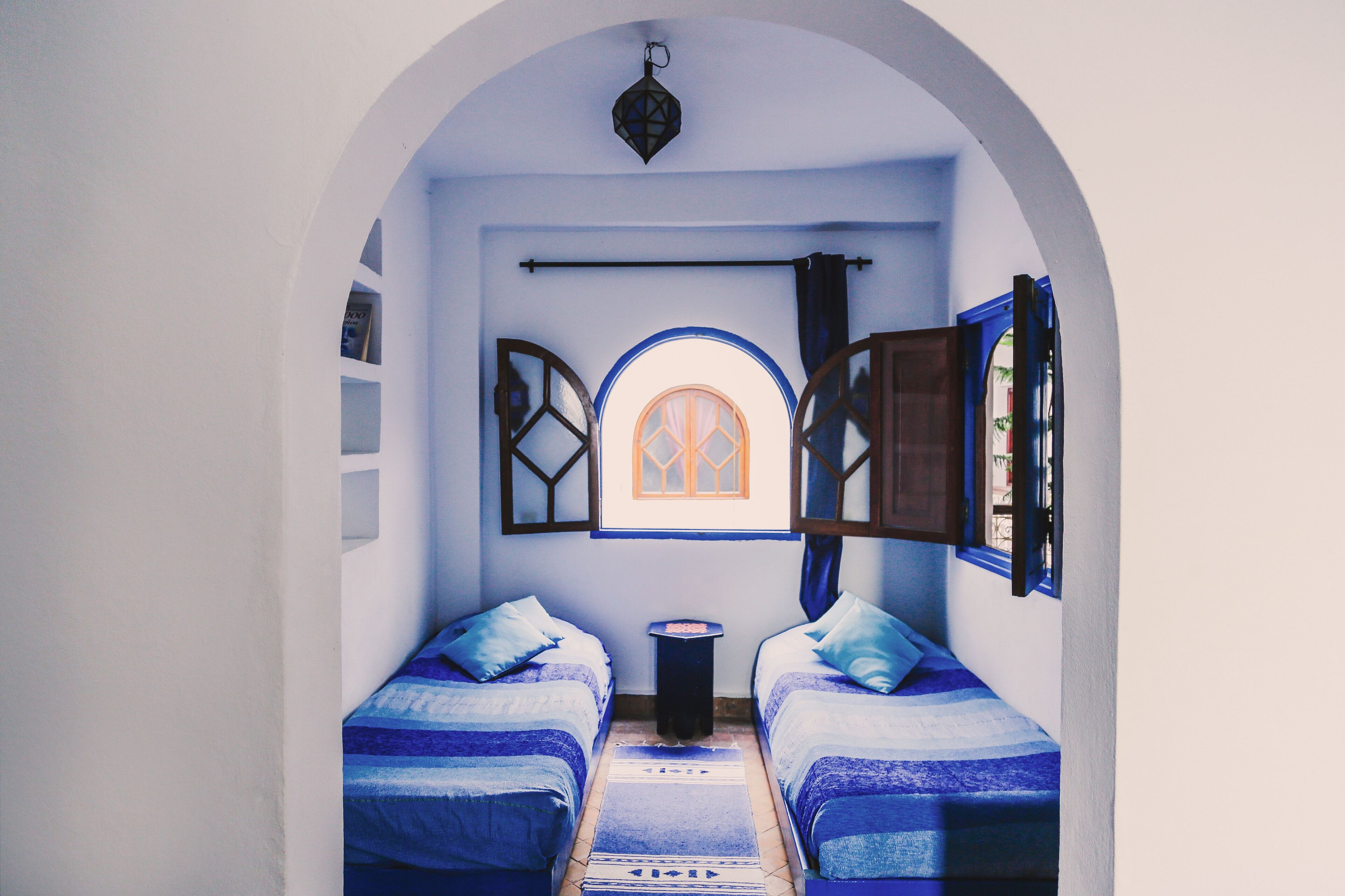 A narrow room with two beds, a small table, and a window with wooden shutters.