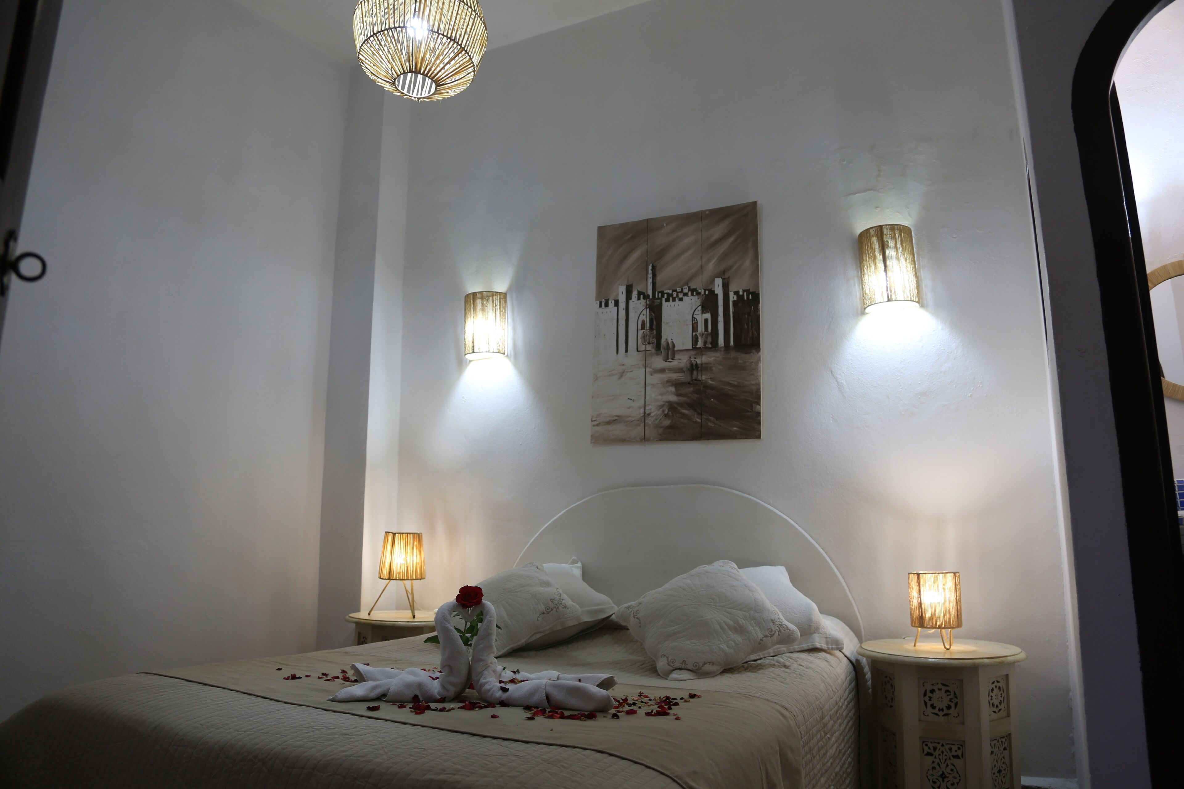 A neatly made bed with white linens, two bedside lamps, a wall-mounted painting, and a decorative light fixture above.