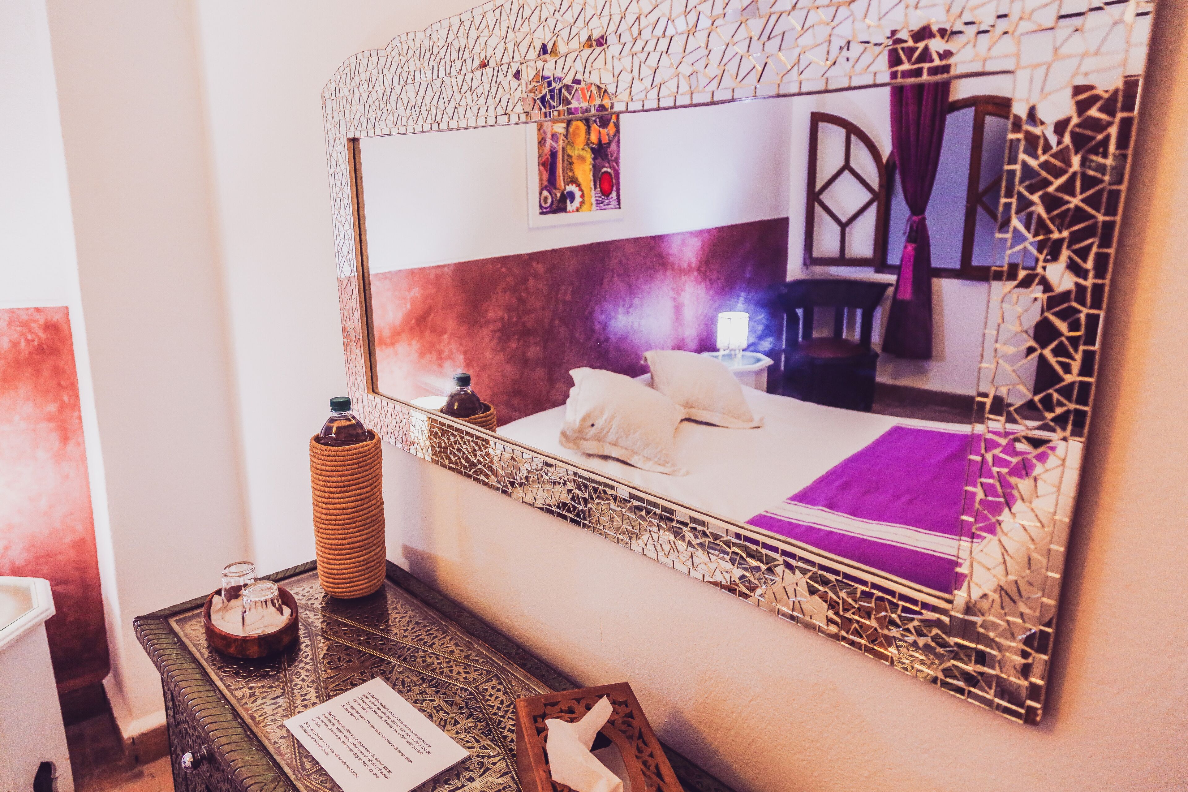 A bedroom with a large mirror, a bed with white linens, a purple throw, and a decorative wall piece.