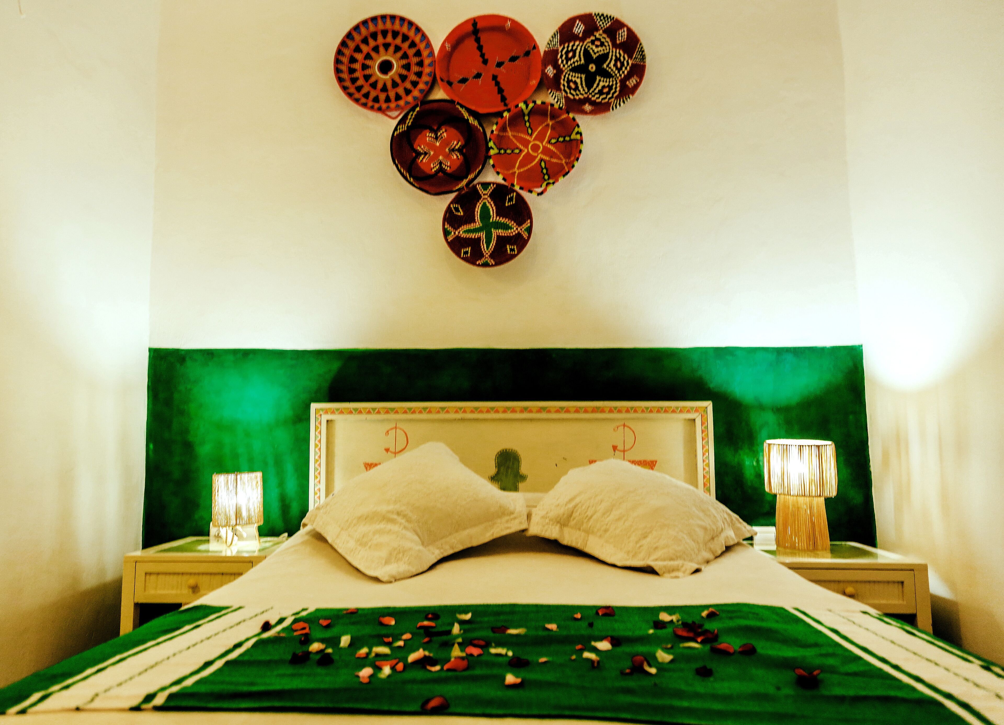 Una cama con cabecera verde, almohadas blancas y colcha verde adornada con pétalos de rosa.